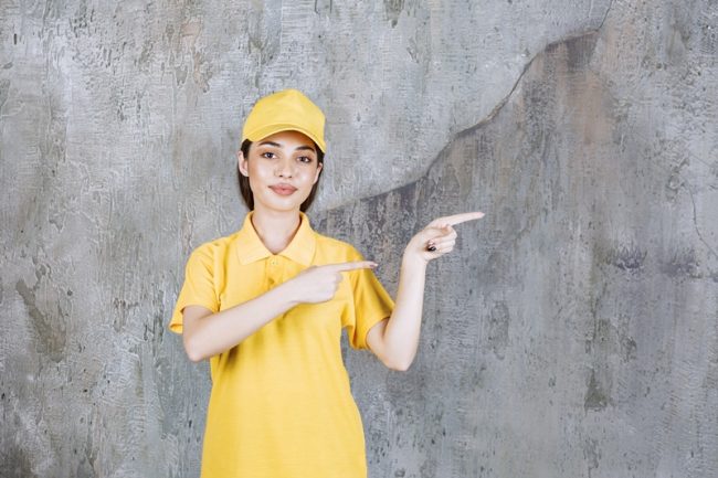 Female service agent in yellow uniform standing on concrete wall background and pointing to the right side